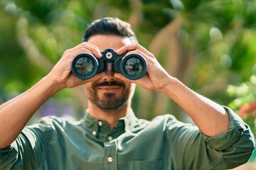Young hispanic man smiling happy looking for new opportunity using binoculars at the park.