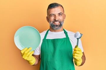Middle age handsome man wearing apron holding scourer washing dishes winking looking at the camera...