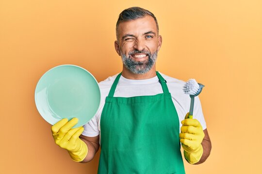 Middle age handsome man wearing apron holding scourer washing dishes winking looking at the camera with sexy expression, cheerful and happy face.