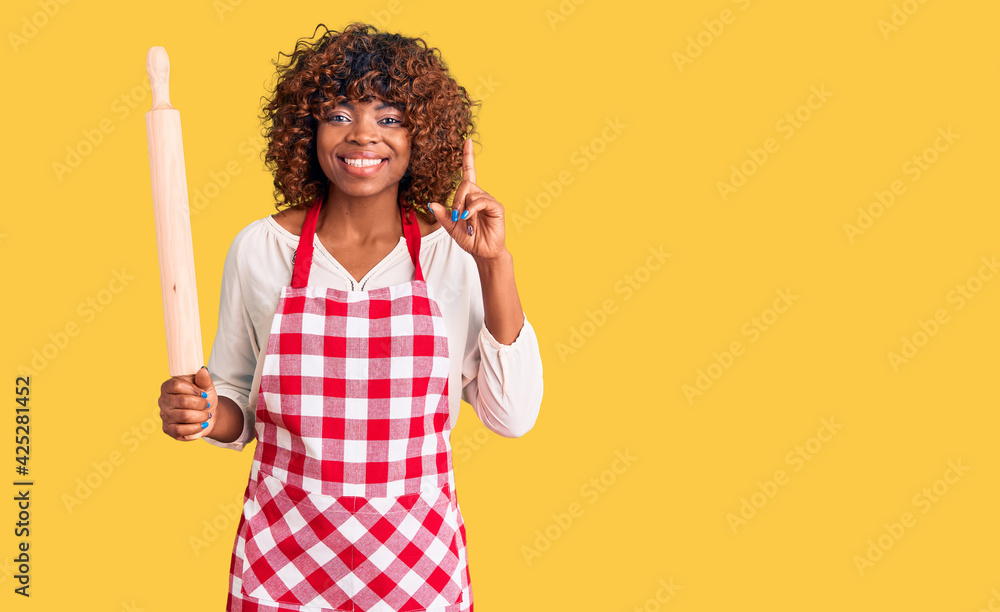 Poster Young african american woman wearing apron holding rolling pin surprised with an idea or question pointing finger with happy face, number one