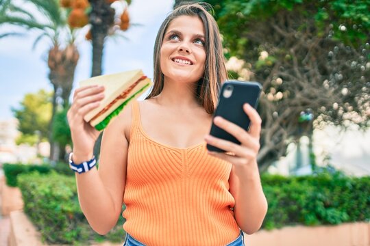 Young middle east girl smiling happy using smartphone and eating sandwich at the city.
