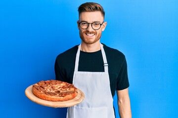 Young redhead man wearing waiter apron holding pizza looking positive and happy standing and smiling with a confident smile showing teeth