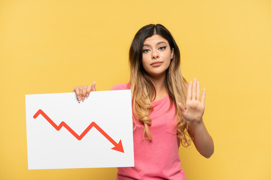 Young Russian girl isolated on yellow background holding a sign with a decreasing statistics arrow symbol and doing stop sign