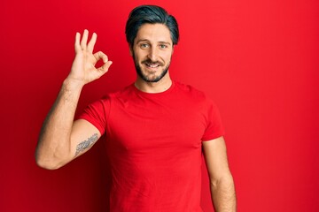 Young hispanic man wearing casual red t shirt smiling positive doing ok sign with hand and fingers....