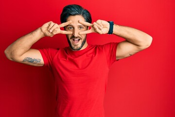 Young hispanic man wearing casual red t shirt doing peace symbol with fingers over face, smiling cheerful showing victory