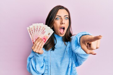 Young brunette woman holding 20 israel shekels banknotes pointing with finger surprised ahead, open...