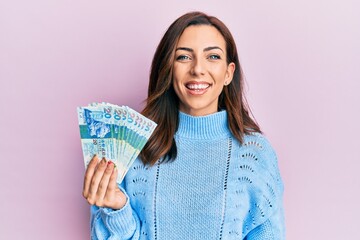 Young brunette woman holding hong kong 20 dollars banknotes looking positive and happy standing and smiling with a confident smile showing teeth