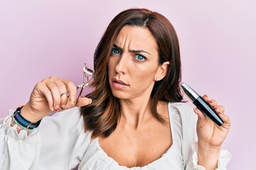 Young brunette woman holding eyelash curler in shock face, looking skeptical and sarcastic,...