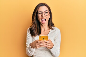 Young brunette woman using smartphone over yellow background winking looking at the camera with...