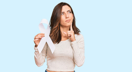 Young brunette woman holding white ribbon serious face thinking about question with hand on chin,...