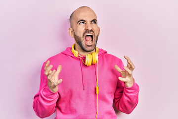 Young bald man wearing gym clothes and using headphones crazy and mad shouting and yelling with aggressive expression and arms raised. frustration concept.
