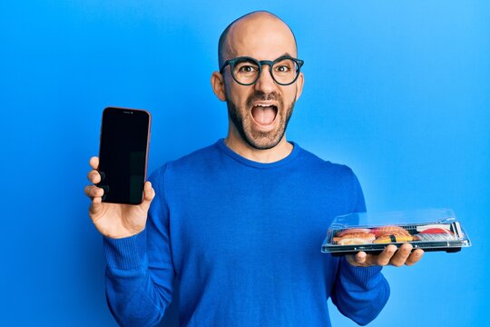 Young hispanic man holding take away food showing smartphone screen celebrating crazy and amazed for success with open eyes screaming excited.
