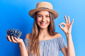 Young beautiful girl wearing summer hat holding bowl with blueberries doing ok sign with fingers,...