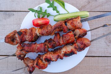 Juicy meat on skewers with tomatoes and herbs on a white plate on a wooden background. Kebab of meat is fried on the grill.