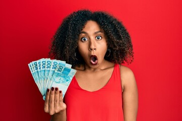 Beautiful african american woman with afro hair holding 100 brazilian real banknotes scared and...