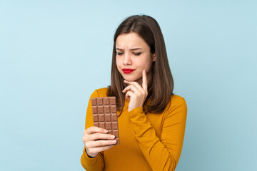 Teenager girl isolated on blue background taking a chocolate tablet and having doubts