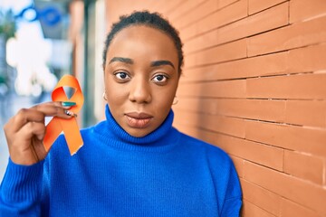 Young african american woman with serious expression holding orange awarenes ribbon at the city.