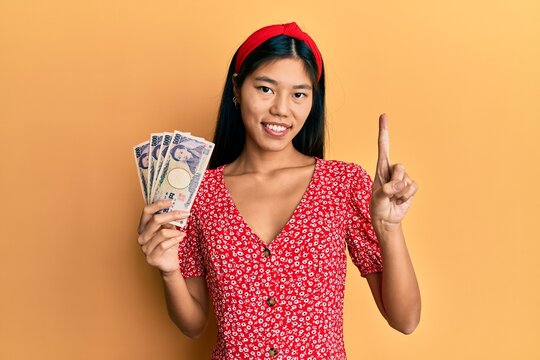 Young chinese woman holding 5000 japanese yen banknotes smiling with an idea or question pointing finger with happy face, number one