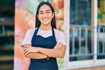 Young latin shopkeeper girl with arms crossed smiling happy at the fruit store. - obrazy, fototapety, plakaty