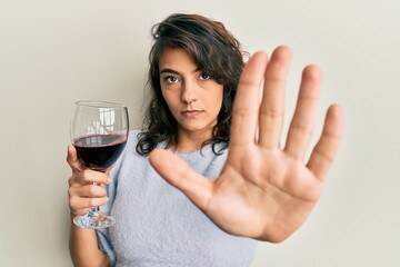 Young hispanic woman drinking a glass of red wine with open hand doing stop sign with serious and confident expression, defense gesture
