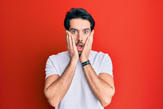 Young hispanic man wearing casual white tshirt afraid and shocked, surprise and amazed expression with hands on face