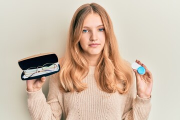 Beautiful young caucasian girl holding glasses and contact lenses relaxed with serious expression...