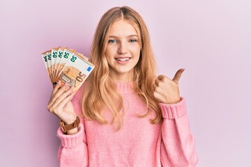 Beautiful young caucasian girl holding bunch of 50 euro banknotes smiling happy and positive, thumb...