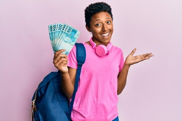 Young african american girl wearing student backpack and holding brazilian real banknotes celebrating achievement with happy smile and winner expression with raised hand
