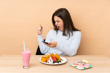 Teenager girl eating waffles isolated on beige background with phone in victory position