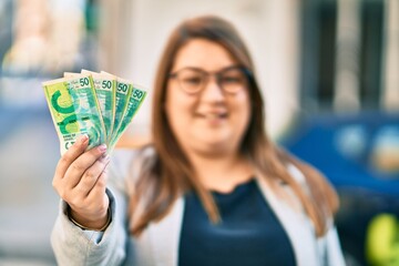 Young hispanic plus size businesswoman smiling happy holding israel shekels banknotes at the city.