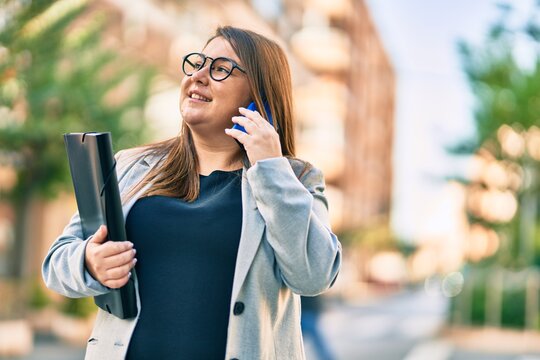 Young plus size businesswoman talking on the smartphone holding binder at the city.