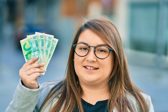 Young hispanic plus size businesswoman smiling happy holding israel shekels banknotes at the city.