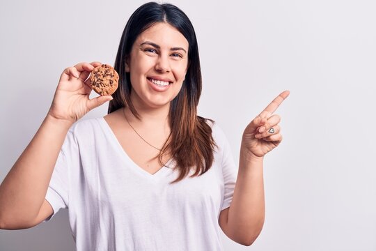 Young beautiful brunette woman eating chocolate cookie over isolated white background smiling happy pointing with hand and finger to the side