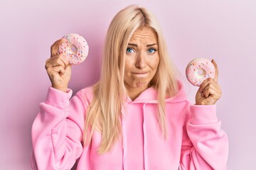 Young blonde woman holding tasty pink doughnuts over eyes skeptic and nervous, frowning upset...