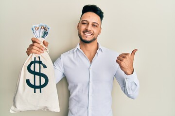 Young arab man holding dollars bag pointing thumb up to the side smiling happy with open mouth