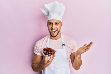 Young arab man wearing professional cook uniform holding bowl with dates celebrating achievement with happy smile and winner expression with raised hand