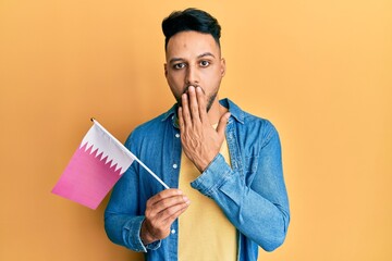 Young arab man holding qatar flag covering mouth with hand, shocked and afraid for mistake....