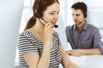 Casual dressed young woman using headset and computer while talking with customers online. Group of operators at work. Call center, business concept