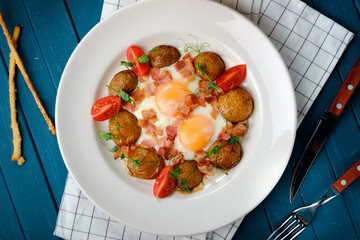 Breakfast with fried eggs with potatoes, bacon and tomatoes on a white plate.
