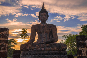 Obraz premium The sculpture of a sitting Buddha against a cloud sunset. The ruins of the ancient Buddhist temple of Wat Chana Songhram. Sukhotai, Thailand