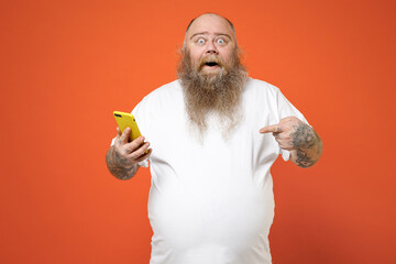 Fat surprised pudge obese chubby overweight tattooed blue-eyed bearded man 30s has big belly in white t-shirt point index finger on mobile cell phone isolated on red orange background studio portrait.