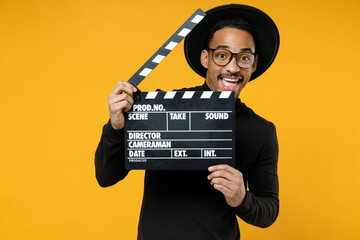 Young happy smiling fun cheerful african man 20s wearing stylish black shirt hat eyeglasses holding classic black film making clapperboard isolated on yellow orange color background studio portrait