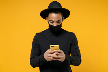 Young african man in trendy stylish black hat shirt face mask to safe from coronavirus covid-19 pandemic quarantine hold mobile phone browsing symptoms isolated on yellow background studio portrait.