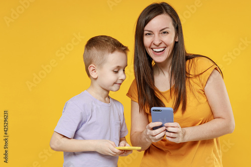 Happy young woman have fun with cute child baby boy 5-6-7 years old in violet t-shirt hold use mobile phone. Mommy little kid son together isolated on yellow background studio Mother's Day love family