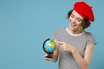 Young geography student teacher woman, short hairdo in french beret red hat striped t-shirt hold in hands spin touch with finger Earth world globe isolated on pastel blue background studio portrait