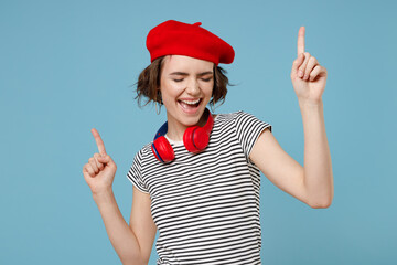 Young fun happy smiling woman 20s with short hairdo wearing french beret red hat striped t-shirt headphones listening music dancing point finger up isolated on pastel blue background studio portrait.