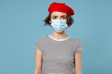 Young woman 20s wearing french beret red hat striped t-shirt sterile face mask to safe from coronavirus virus covid-19 during pandemic quarantine isolated on pastel blue background studio portrait.