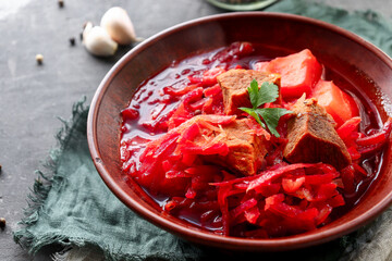 Red borsch with vegetables and meat in a clay plate. Steam from hot tomato soup. Dark background. Delicious healthy lunch. Copy space