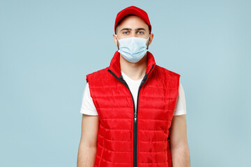 Delivery guy employee man in red cap white t-shirt vest waistcoat uniform sterile face mask gloves work courier service on lockdown coronavirus virus looking camera isolated on pastel blue background.