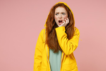 Redhead young puzzled confused woman in yellow waterproof raincoat outerwear put on hood look aside biting nails fingers isolated on pastel pink background studio Outdoors wet weather fall concept